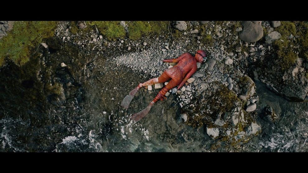 פריים מתוך הסרט סלמון פראי, דמות חצי אדם חצי דג סלמון שוכבת על הבטן על סלע בתוך נהר